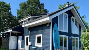 Una casa de dos pisos con fachada azul, una terraza de madera y una tabla de surf apoyada contra la casa.