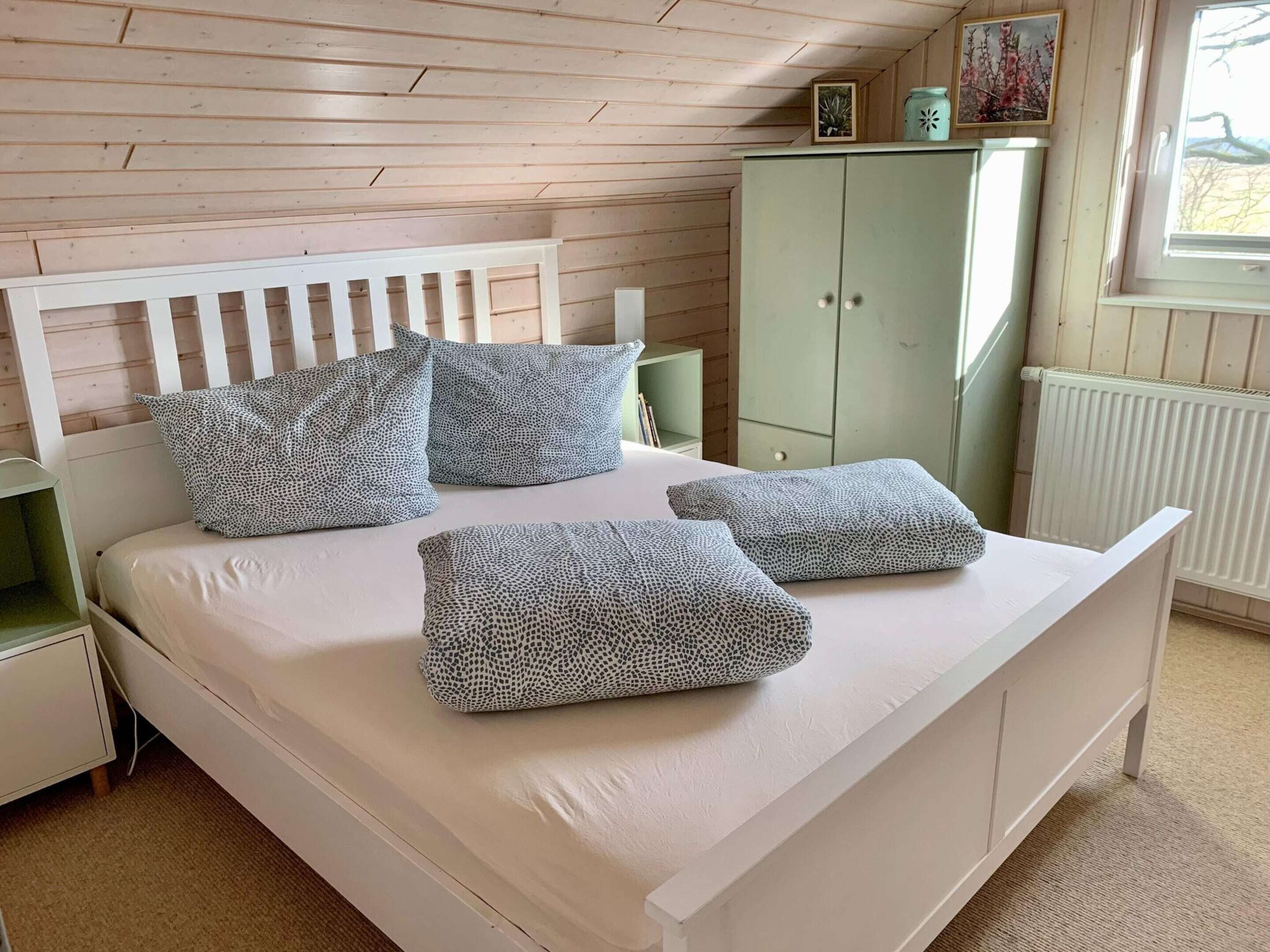 A bedroom with a white bed, patterned pillows, a green wardrobe, and a window with a view.
