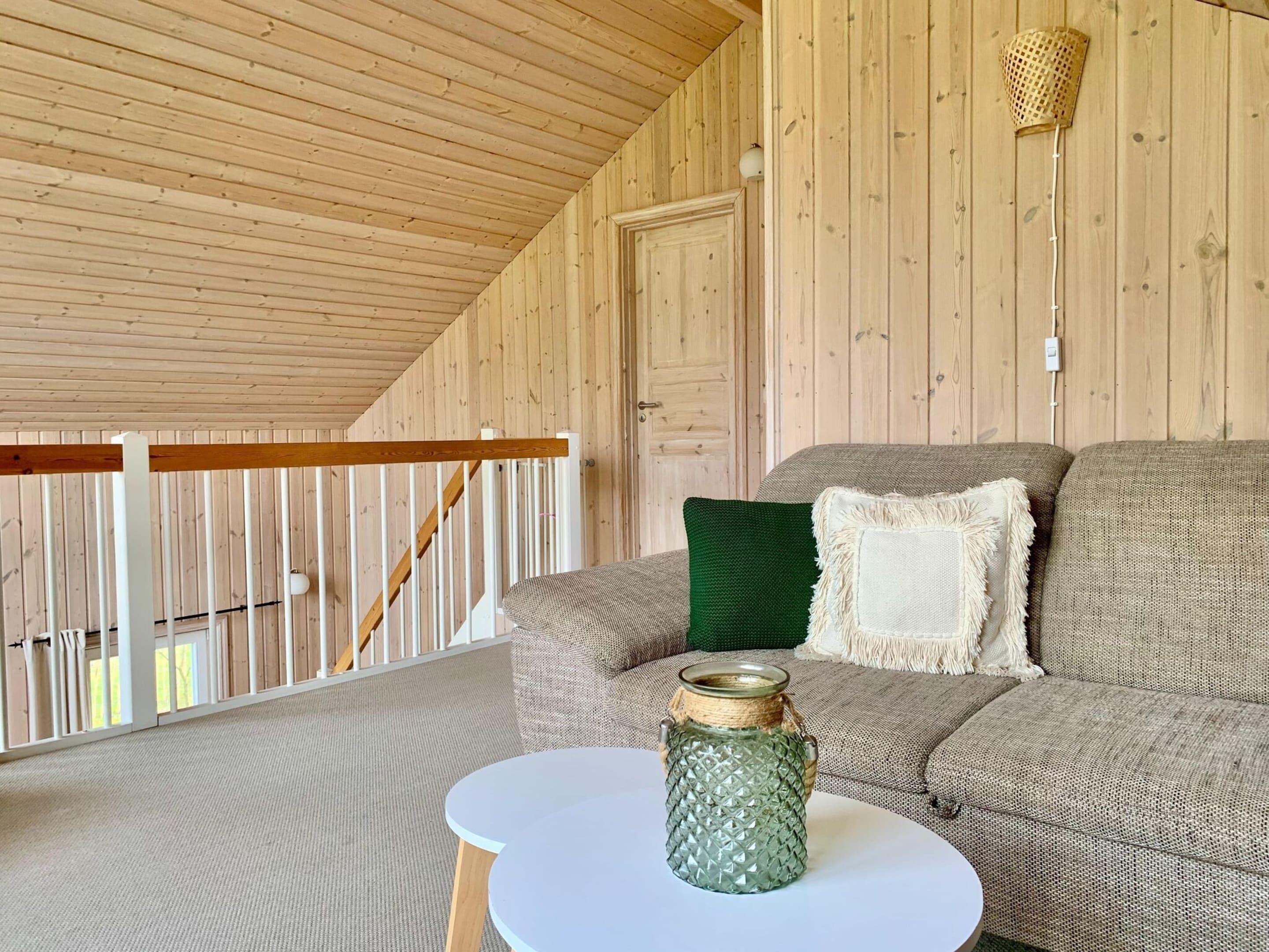 A cozy room with a wooden ceiling, a white railing, a grey sofa with a green pillow, and a small table with a glass jar.