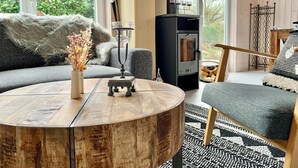Una sala de estar acogedora con una mesa de centro redonda de madera, un sofá con cojines suaves y un estufa de leña negra, y vistas al verdor exterior.