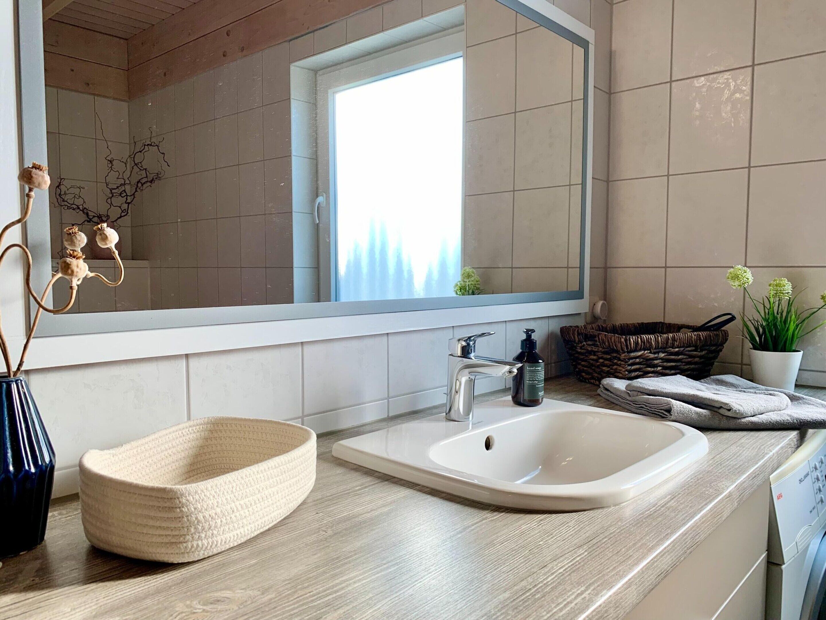 Een moderne badkamer met een grote spiegel, een wastafel, een houten aanrechtblad, een gevlochten mand, een potplant en een droogrek.