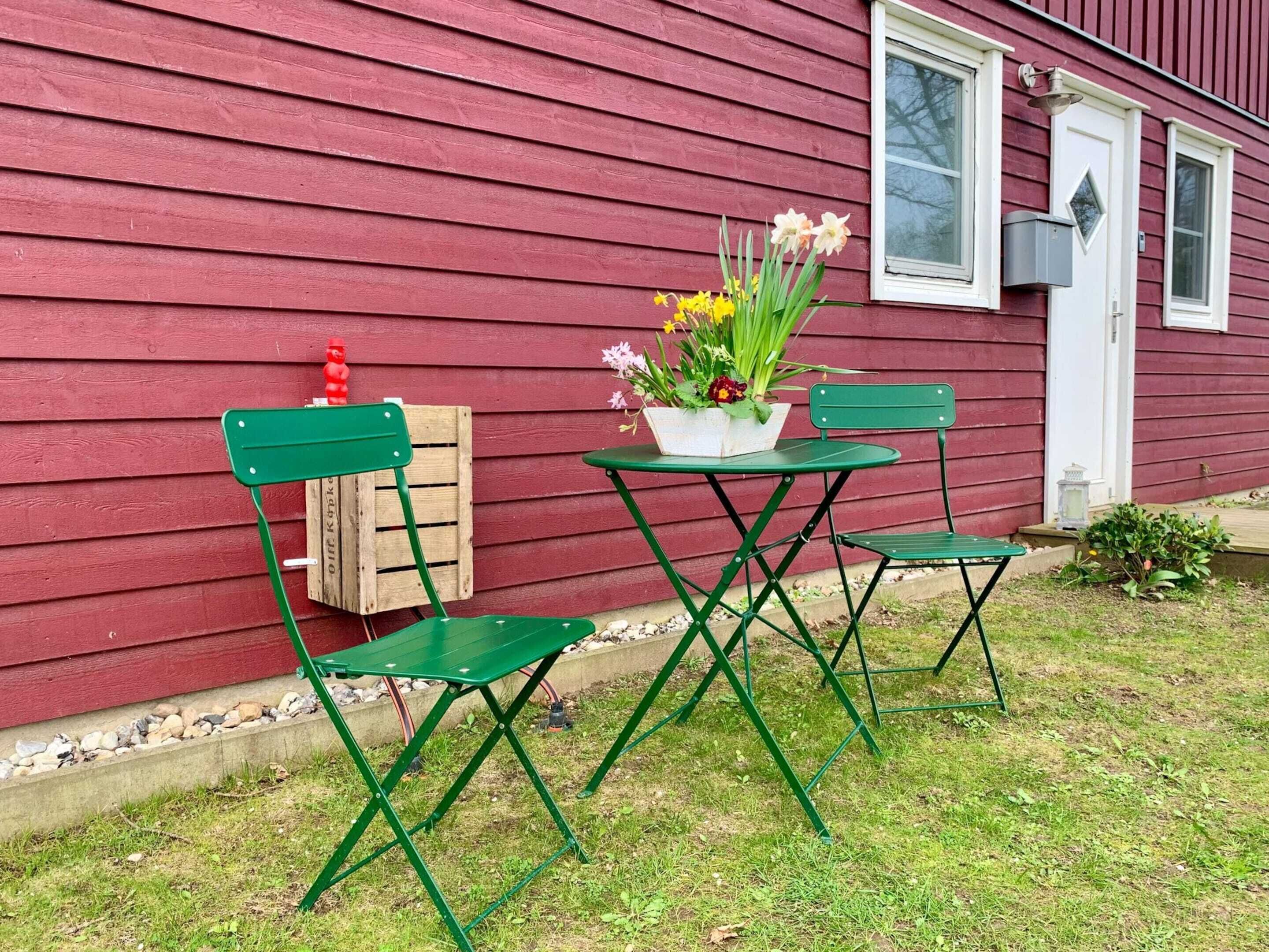 Una casa de madera roja con una puerta y una ventana blancas, una mesa plegable verde con dos sillas, y una maceta con flores amarillas en el porche.