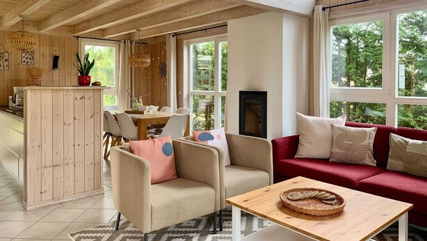 Amplia sala de estar con techo alto, vigas de madera, una mesa de comedor con sillas, un sofá rojo y grandes ventanales que ofrecen vistas a la vegetación.