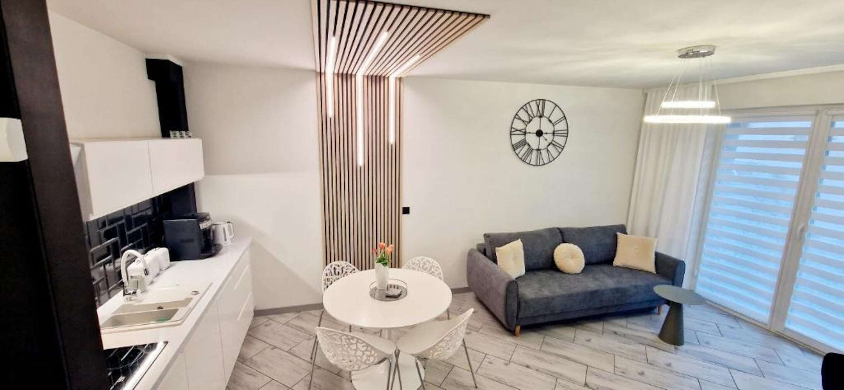 A modern apartment with a kitchen, dining area, and living room. It features a white kitchen with built-in cabinets, a dining table with chairs, a gray sofa with cushions, and a large window with blinds.