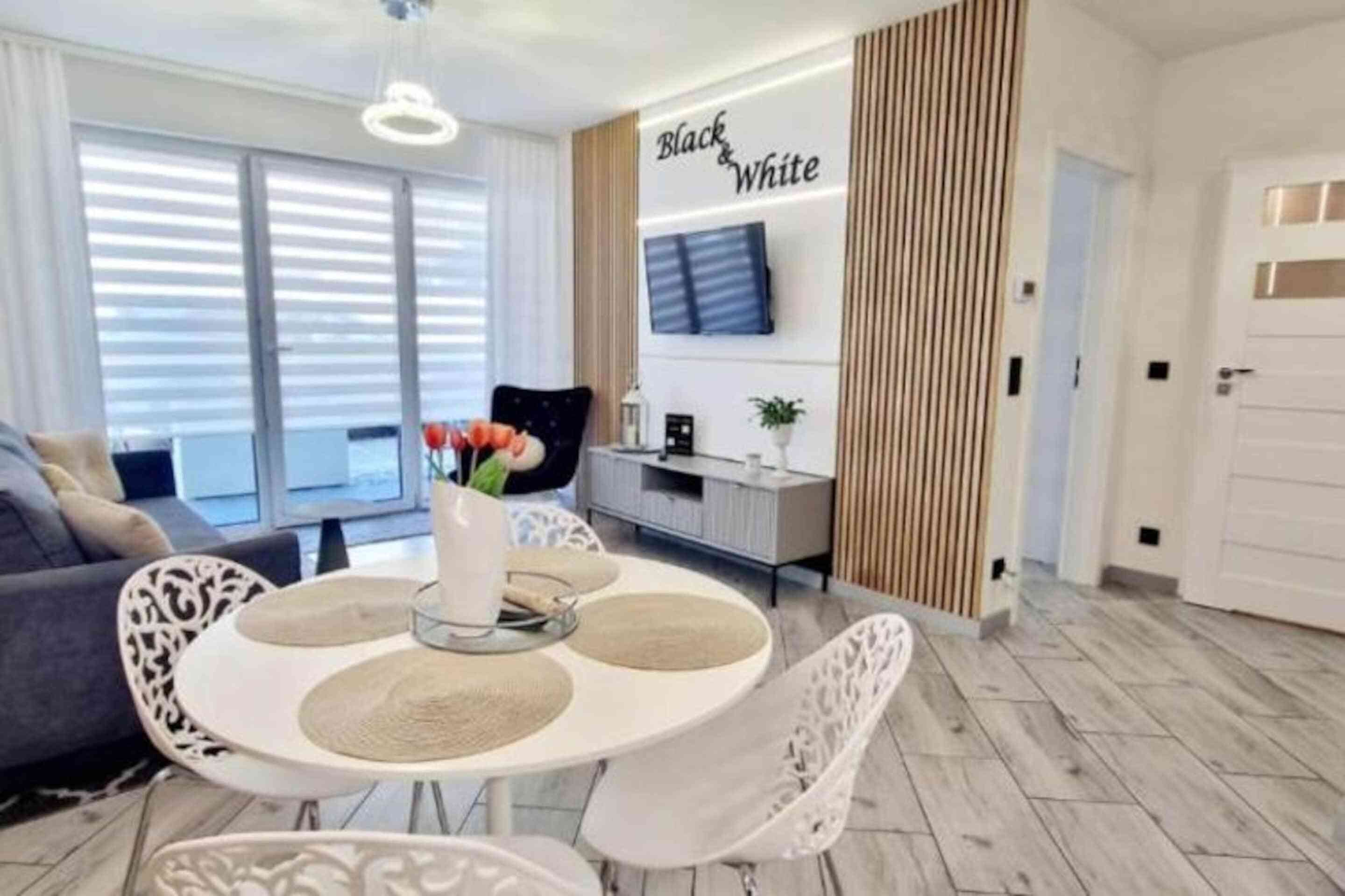 Una sala de estar moderna con una mesa de comedor redonda, sillas blancas y un sofá. Un televisor de pantalla plana está montado en la pared, y hay un letrero decorativo que dice 'Black & White'.