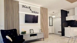 A modern living room with a black and white color scheme, a flat-screen TV mounted on the wall, a black chair, a white chair, and a kitchen area in the background. - Apartment With a Terrace in Mielno (Mielno)