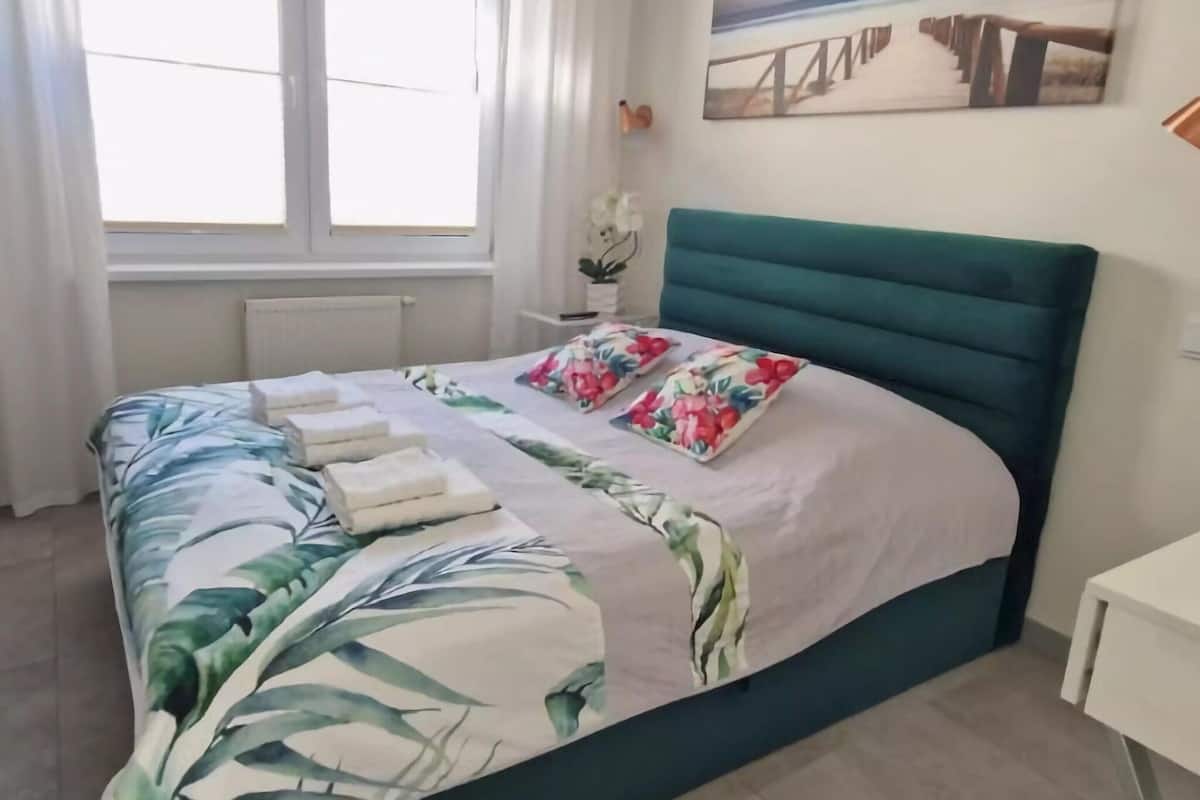 A neatly made bed with floral bedding, a teal headboard, and a painting of a bridge on the wall.