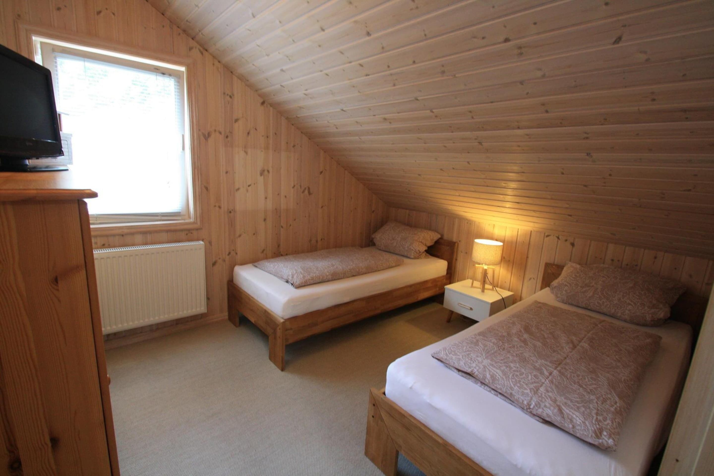 A small, cozy attic bedroom with two single beds, a nightstand, a lamp, and a television on a wooden cabinet.