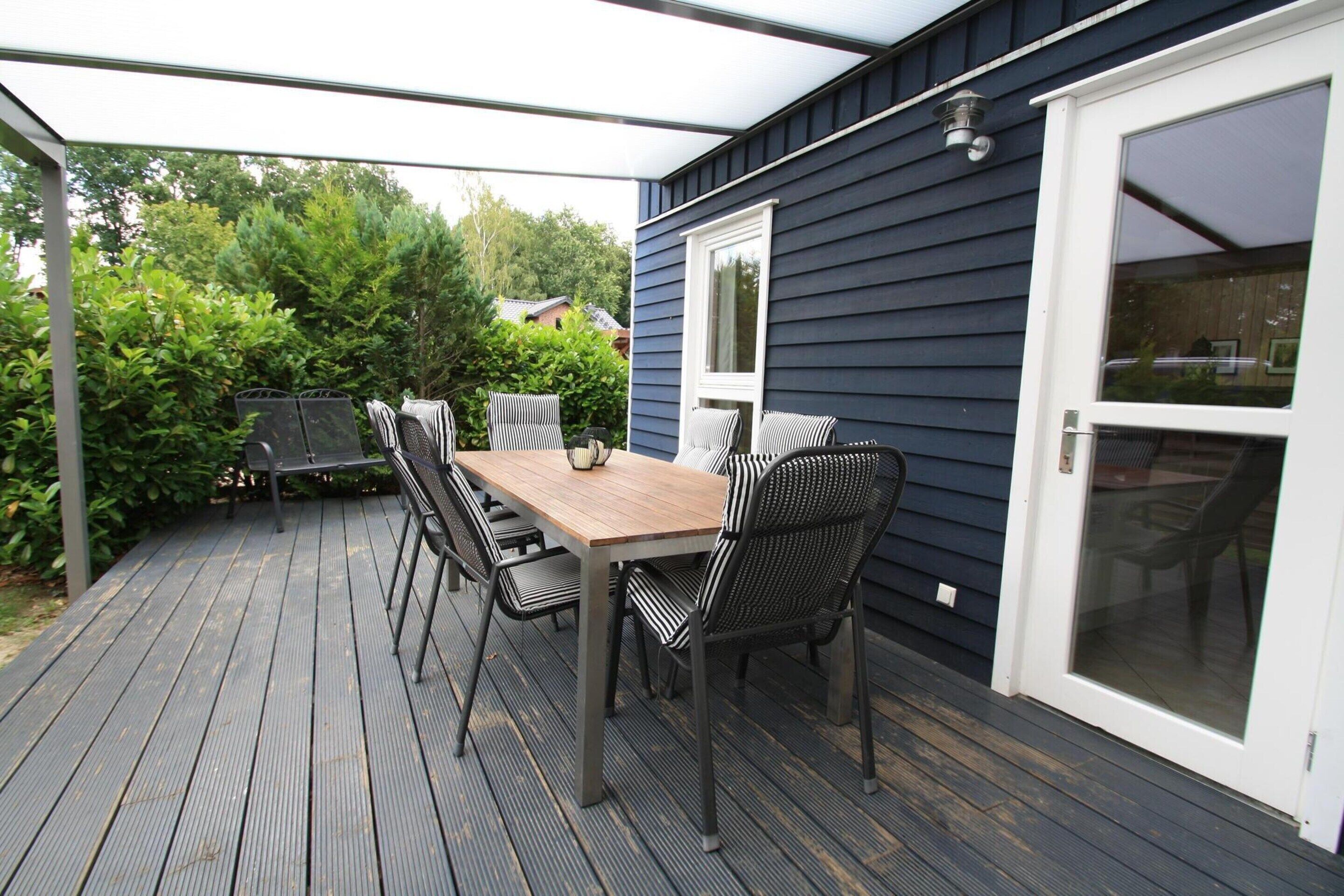 A covered patio with a wooden table and chairs, a sliding glass door, and a view of greenery.