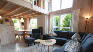 Un vaste espace de vie doté d’un plafond haut, d’un canapé de couleur sombre agrémenté de coussins à motifs, d’une table basse en bois et d’un coin repas avec une table et des chaises blanches.