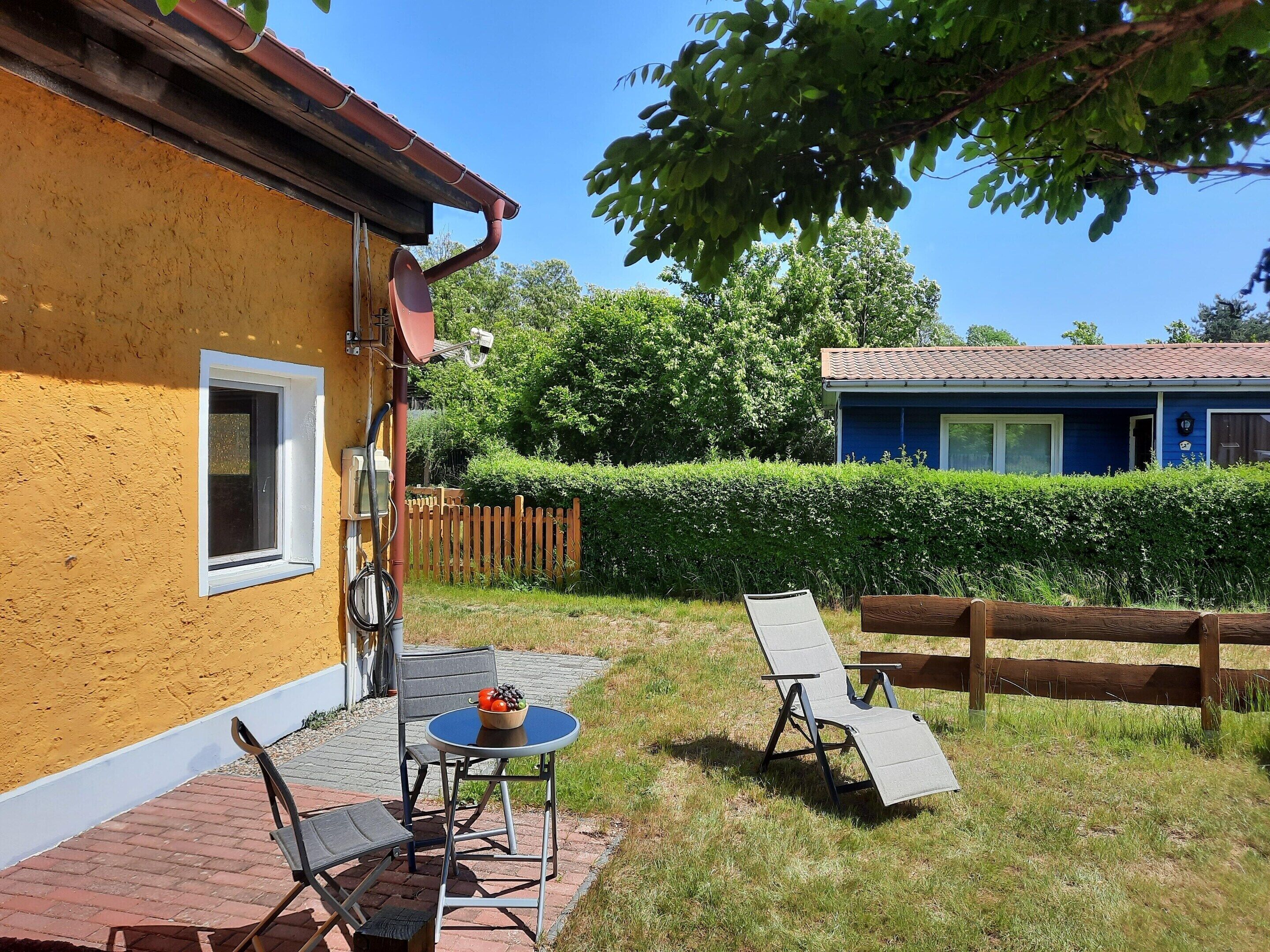 Un espace patio aménagé avec du mobilier d’extérieur, une petite table sur laquelle se trouve un saladier rempli de fruits, et une vue sur une maison bleue avec un toit en tuiles.