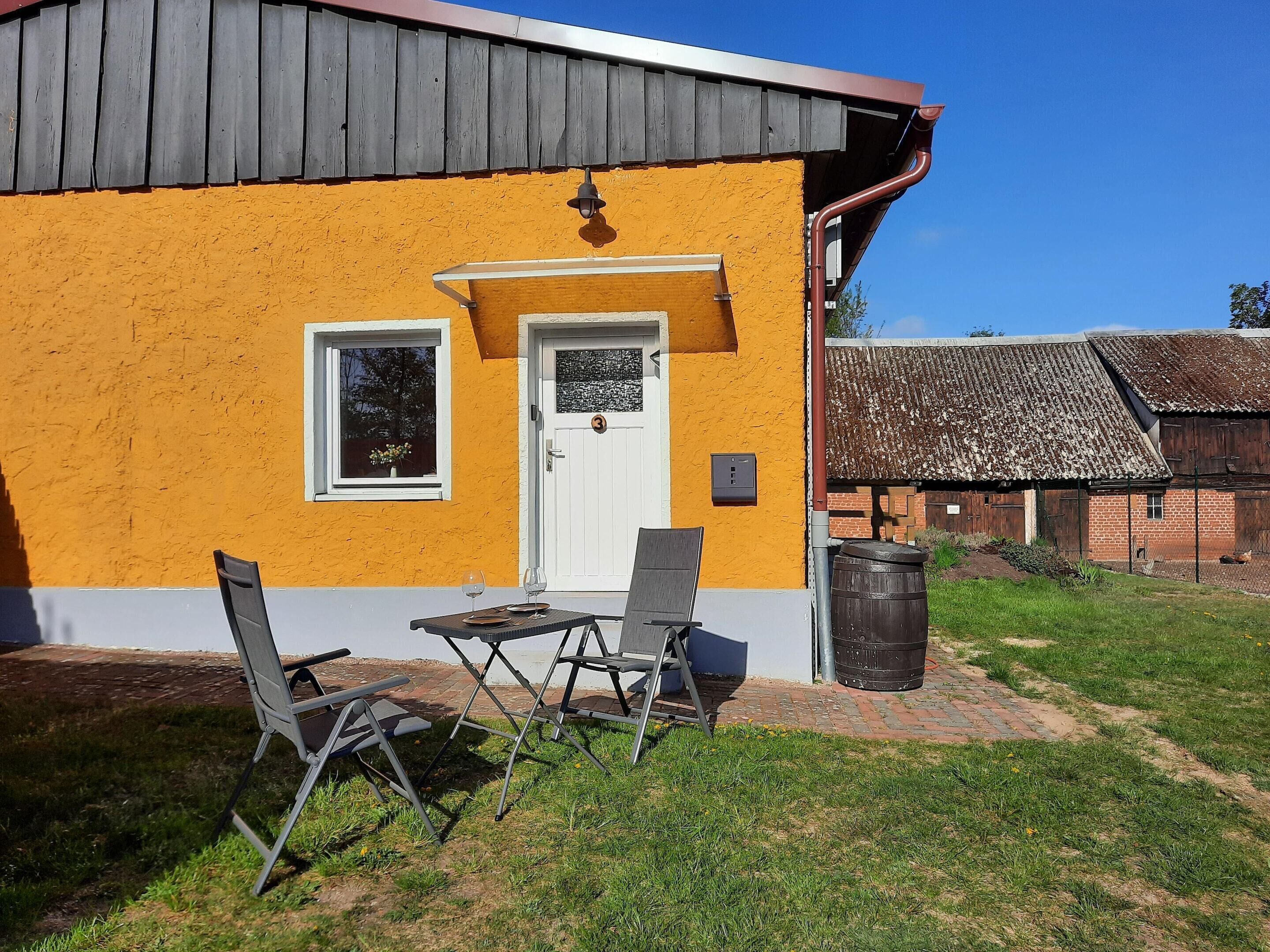 A yellow building with a white door and window, a small table with chairs, and a brick pathway leading to it.