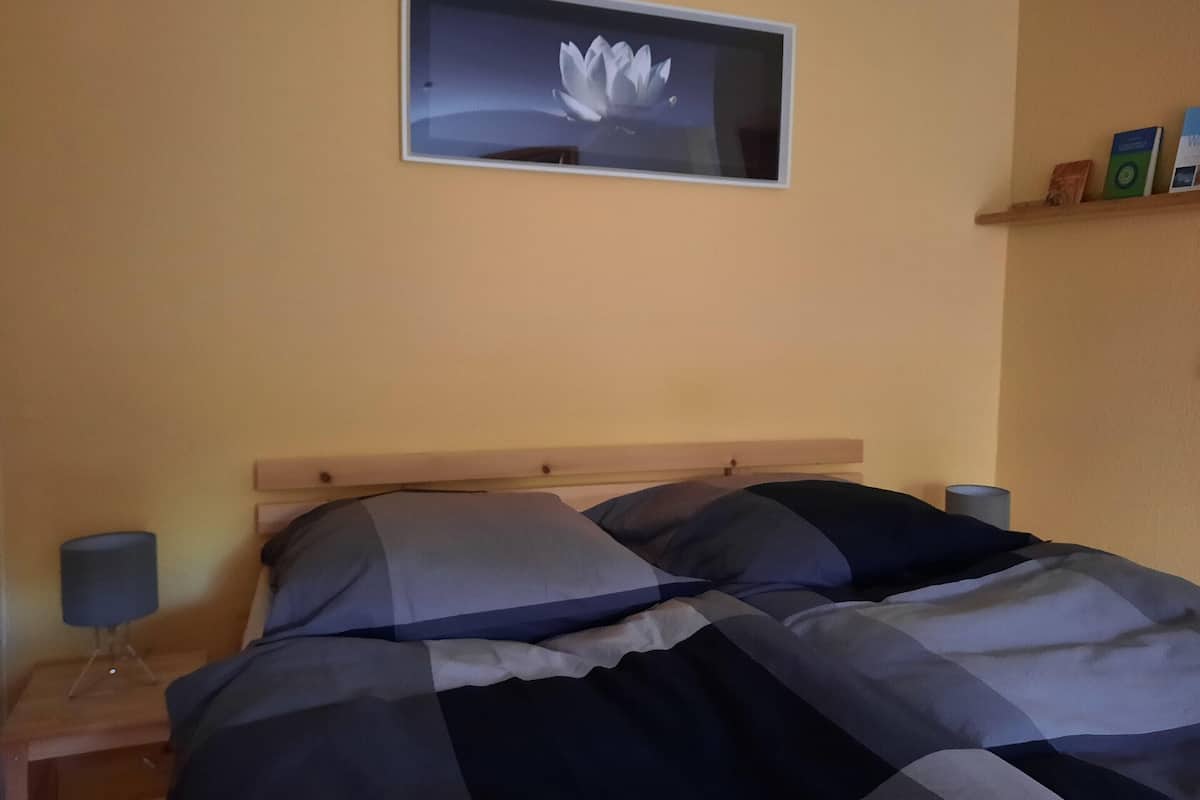 A bed with a wooden headboard, a wall-mounted picture of a lotus flower, and a shelf with books and a lamp.