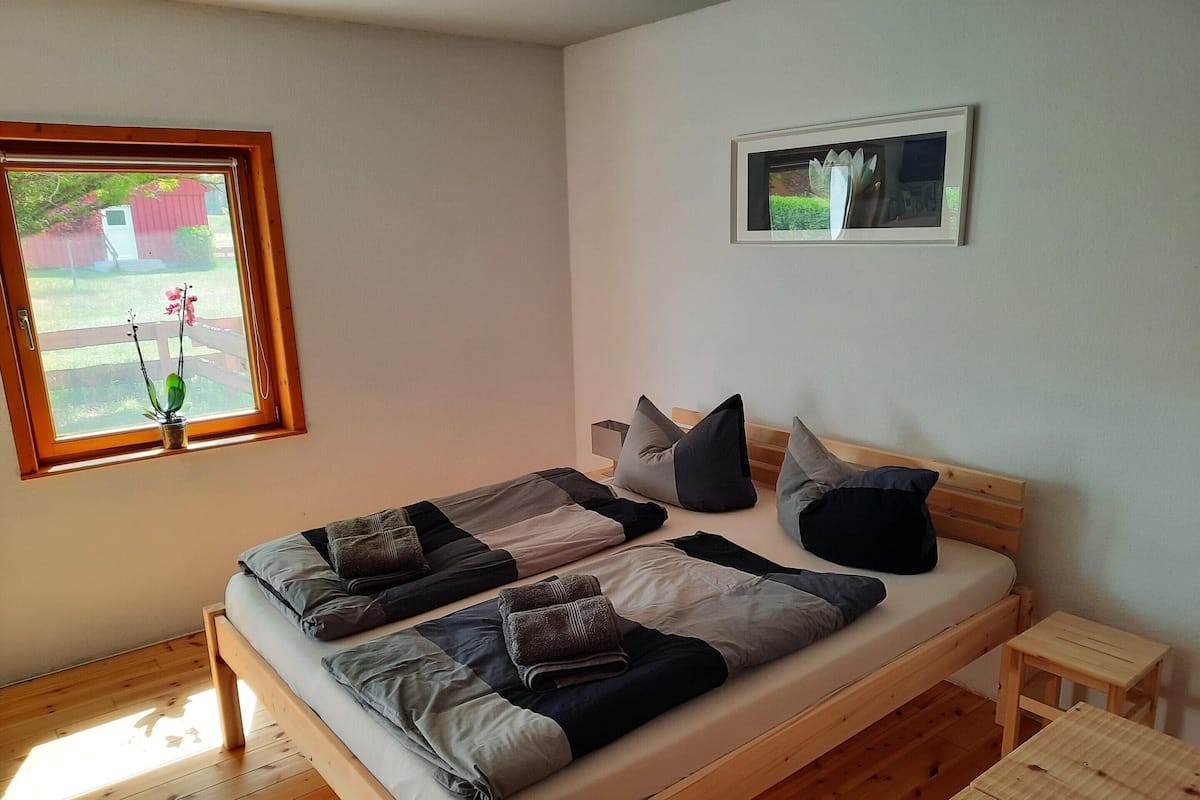 A bedroom with a wooden bed, pillows, a small bedside table, a window with a view of a garden, and a framed picture on the wall.