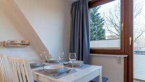 A dining area with a white table set for four, a wooden chair, a window with curtains, and a view of trees outside. - Comfortable Holiday Apartment in St. Peter Ording (St. Peter-Ording)