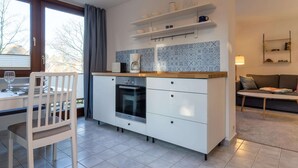 A modern kitchen with white cabinets, a built-in oven, and a coffee maker. There is a dining area with a white table and chair, and a sofa in the living area. - Comfortable Holiday Apartment in St. Peter Ording (St. Peter-Ording)
