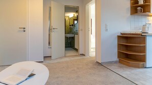A modern hotel room with a white door, a small reading area with a book, a kitchen area with a coffee maker, and a bathroom visible through an open door. - Comfortable Holiday Apartment in St. Peter Ording (St. Peter-Ording)
