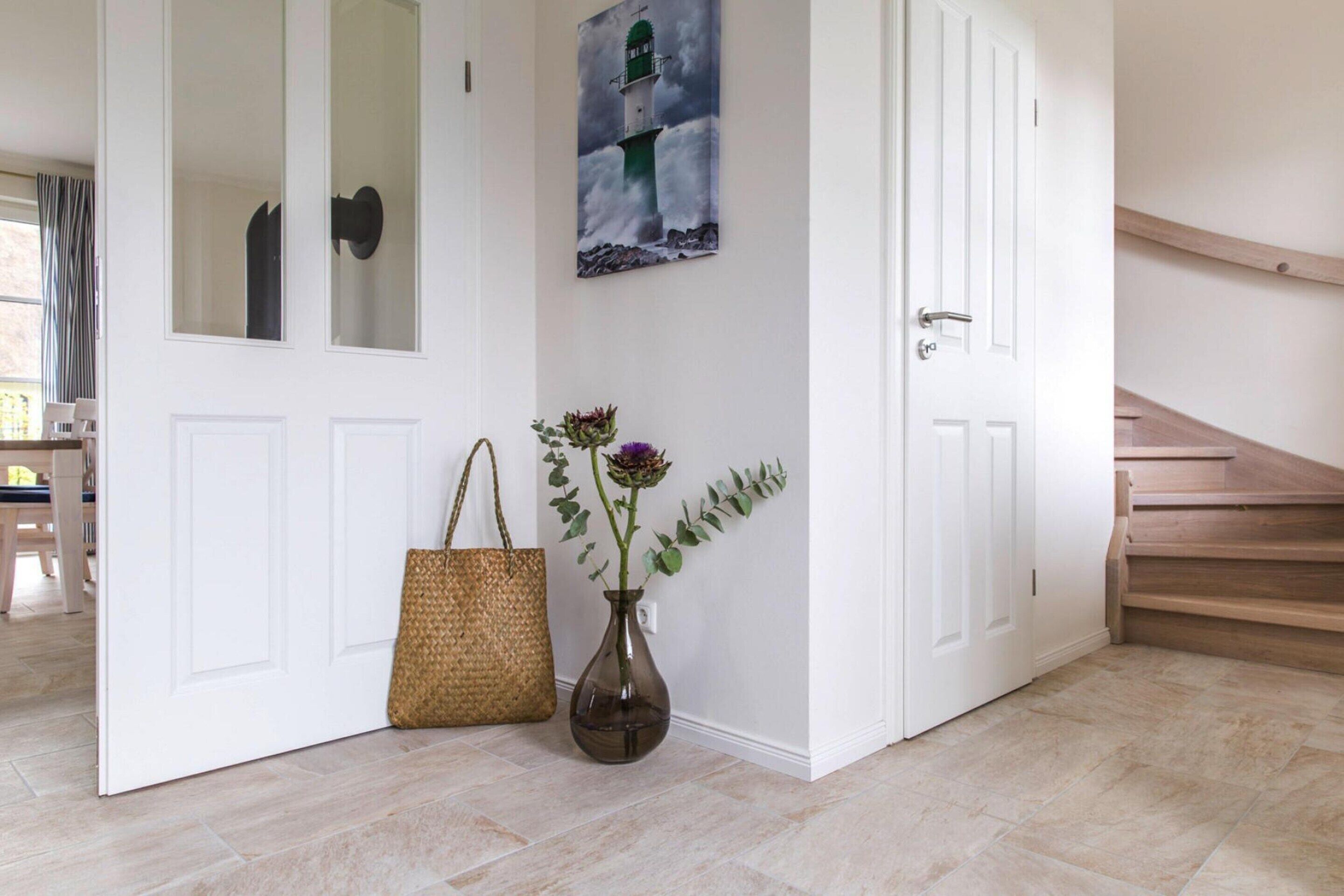 A hallway with a white door, a painting of a lighthouse, a wicker basket, and a vase with flowers.