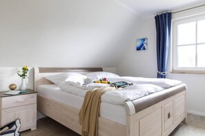 A neatly made bed with a beige blanket, a bedside table with a vase of flowers, and a window with blue curtains. - Holiday House, Fuhlendorf (Fuhlendorf)