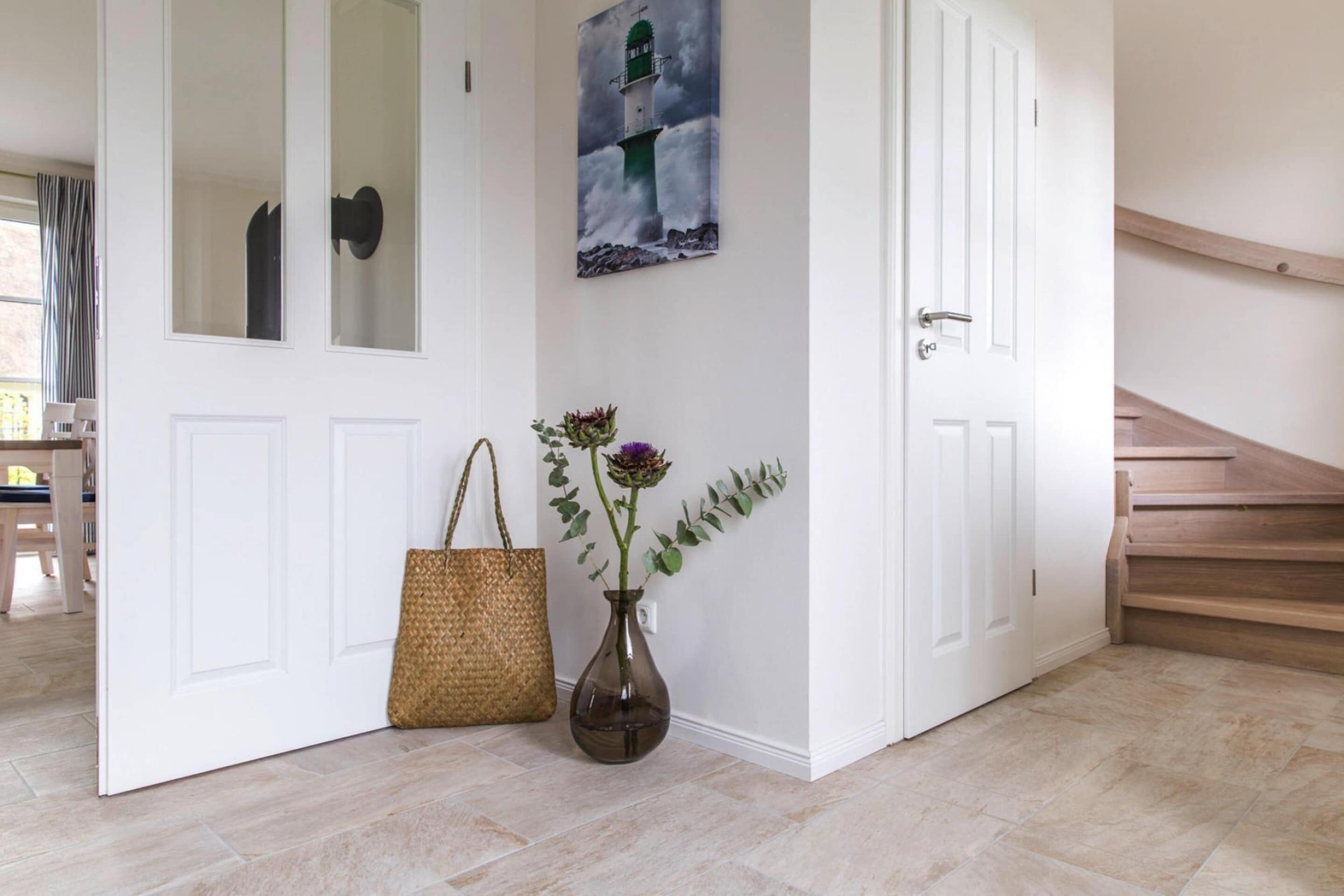 Un pasillo con una puerta blanca, un bolso tejido y un florero con flores.