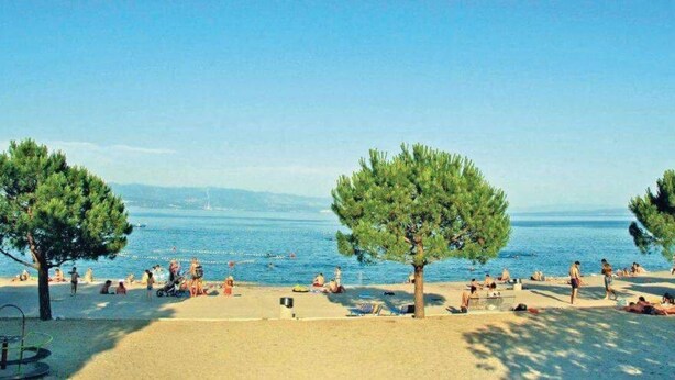 Μια παραλία γεμάτη κόσμο που κάνει ηλιοθεραπεία και κολυμπά, περιτριγυρισμένη από δέντρα και με έναν καθαρό, γαλάζιο ουρανό.