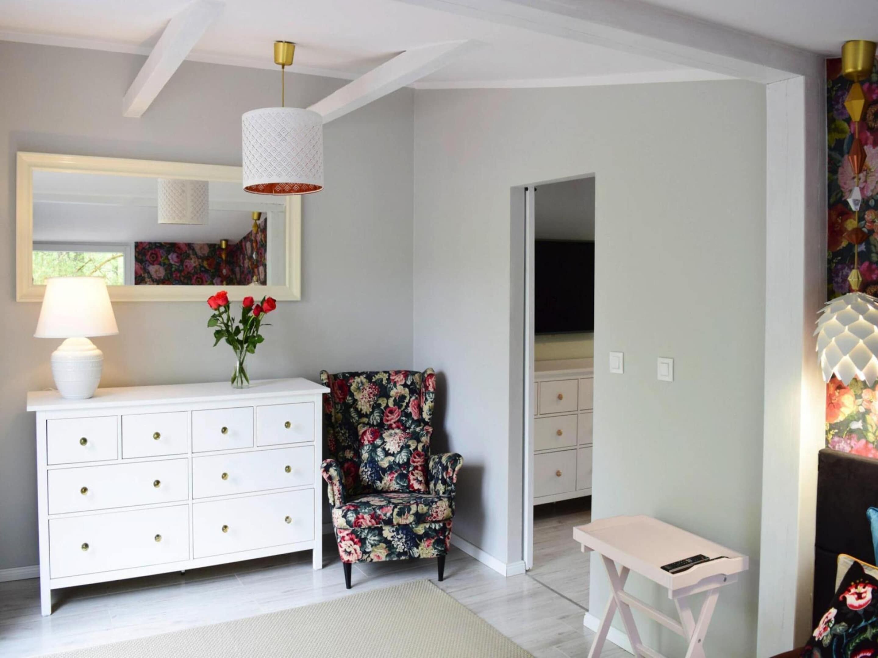 A room with a white dresser, a floral armchair, a small white table, and a hanging light fixture.