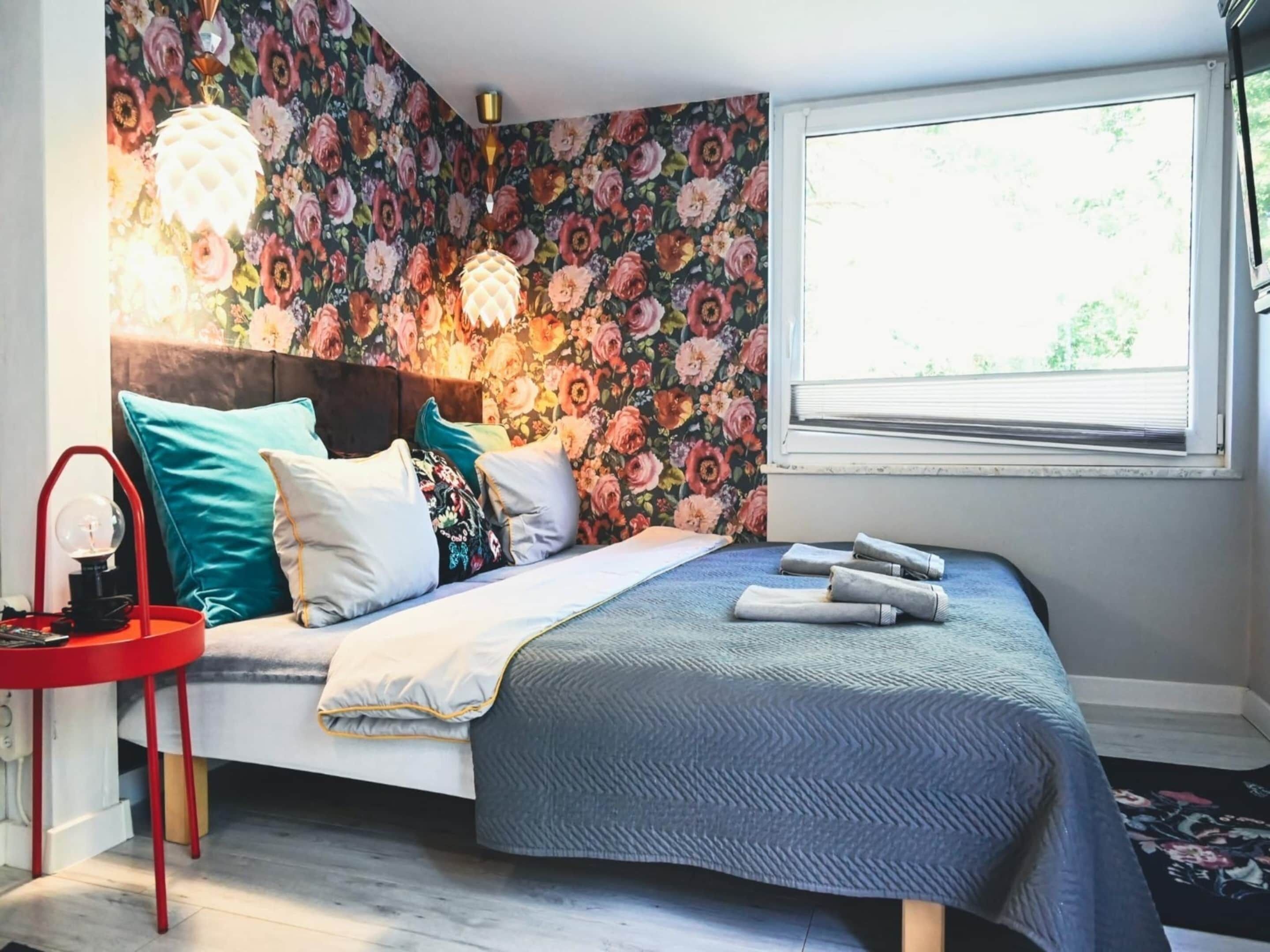 A bedroom with a floral wallpaper, a bed with multiple pillows, a red side table, and a window with blinds.
