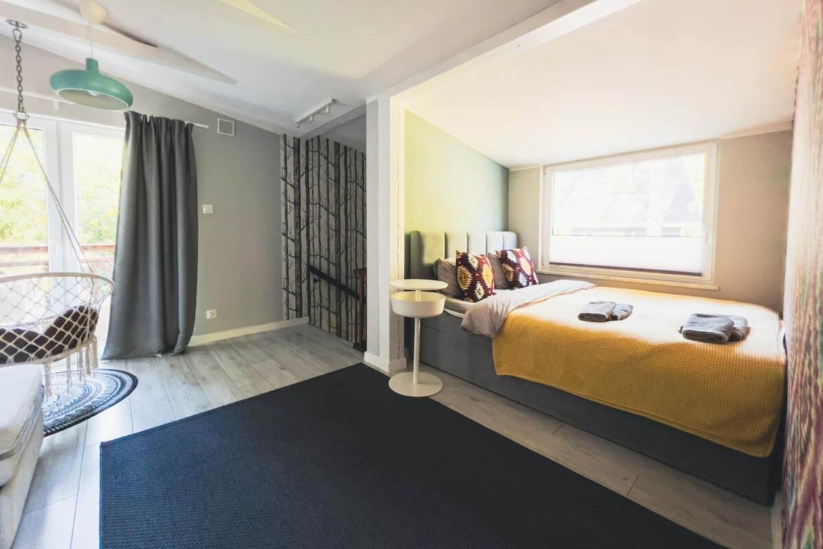 Une chambre à coucher de taille réduite, comprenant un lit, un fauteuil suspendu, une petite table de chevet et une fenêtre.