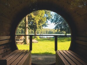 Ein rundes Holzfenster mit einer Bank im Inneren, das auf eine Wiese mit Bäumen und einem Gewässer blickt.