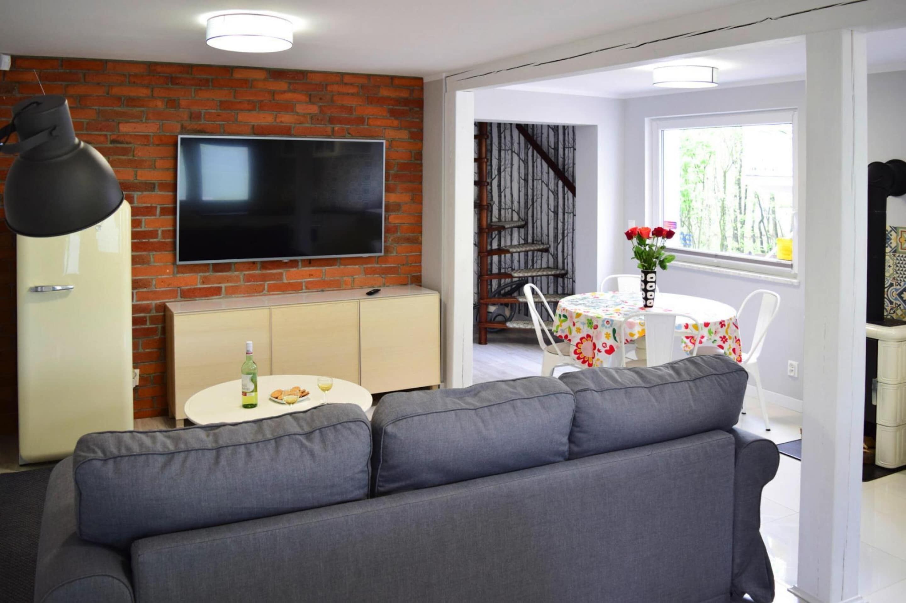 A modern living room with a grey sofa, a TV mounted on a brick wall, a small dining table with a floral tablecloth, and a staircase leading to an upper level.