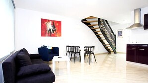 A modern living room with a dark sofa, a blue armchair, and a white coffee table. A staircase with a wooden railing leads to a kitchen area with dark cabinets.