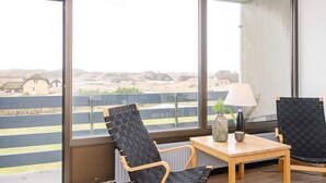 Ein Zimmer mit großen Fenstern, einem Holztisch, zwei Stühlen, einer Lampe und Blick auf Häuser und eine Straße.
