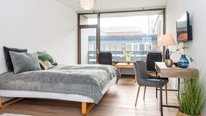 Ein modernes Schlafzimmer mit Bett, Schreibtisch und Stuhl. Ein großes Fenster bietet Blick auf Gebäude.