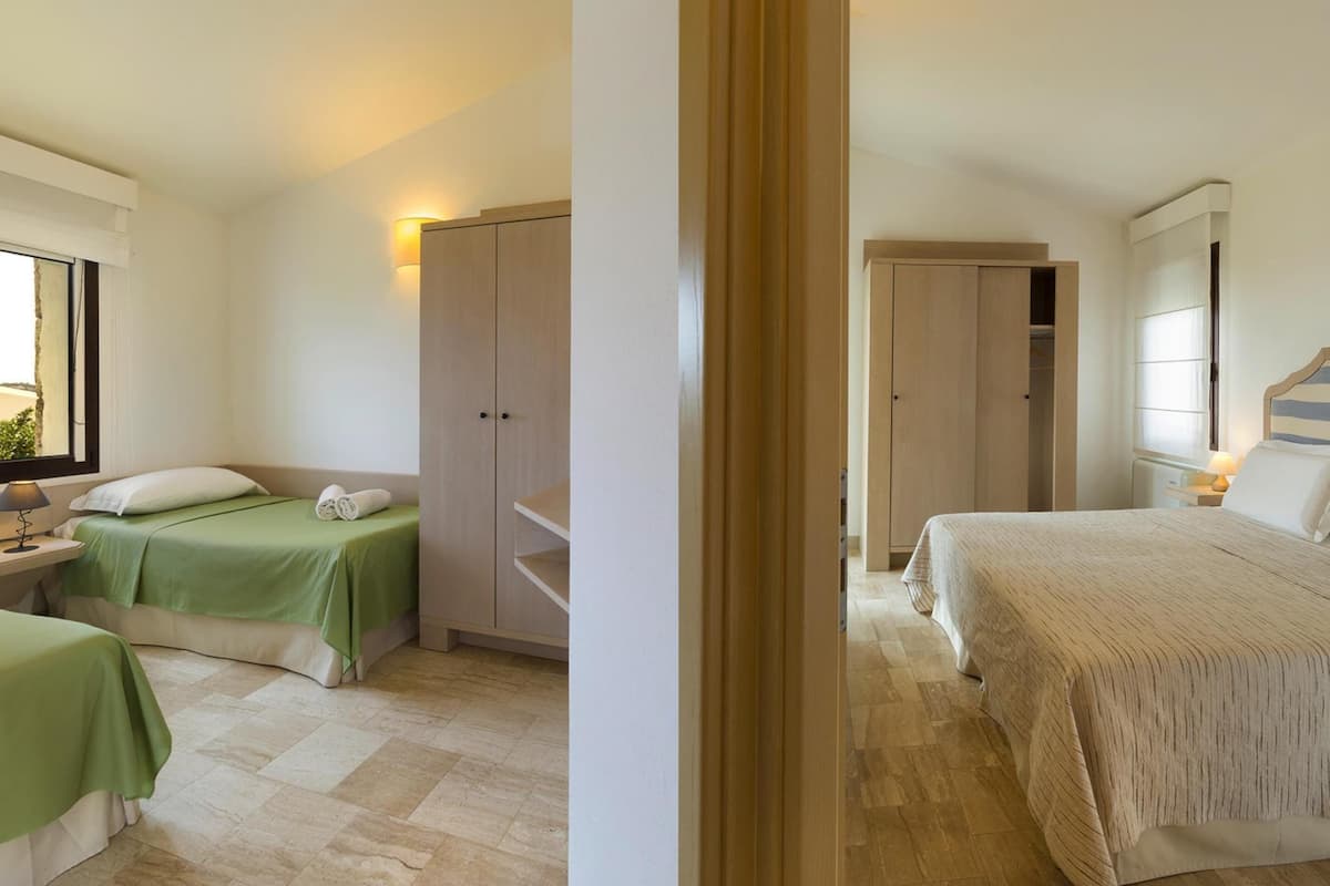 Quarto de hotel com duas camas, um guarda-roupa, uma mesa de cabeceira com abajur e uma janela com cortinas.