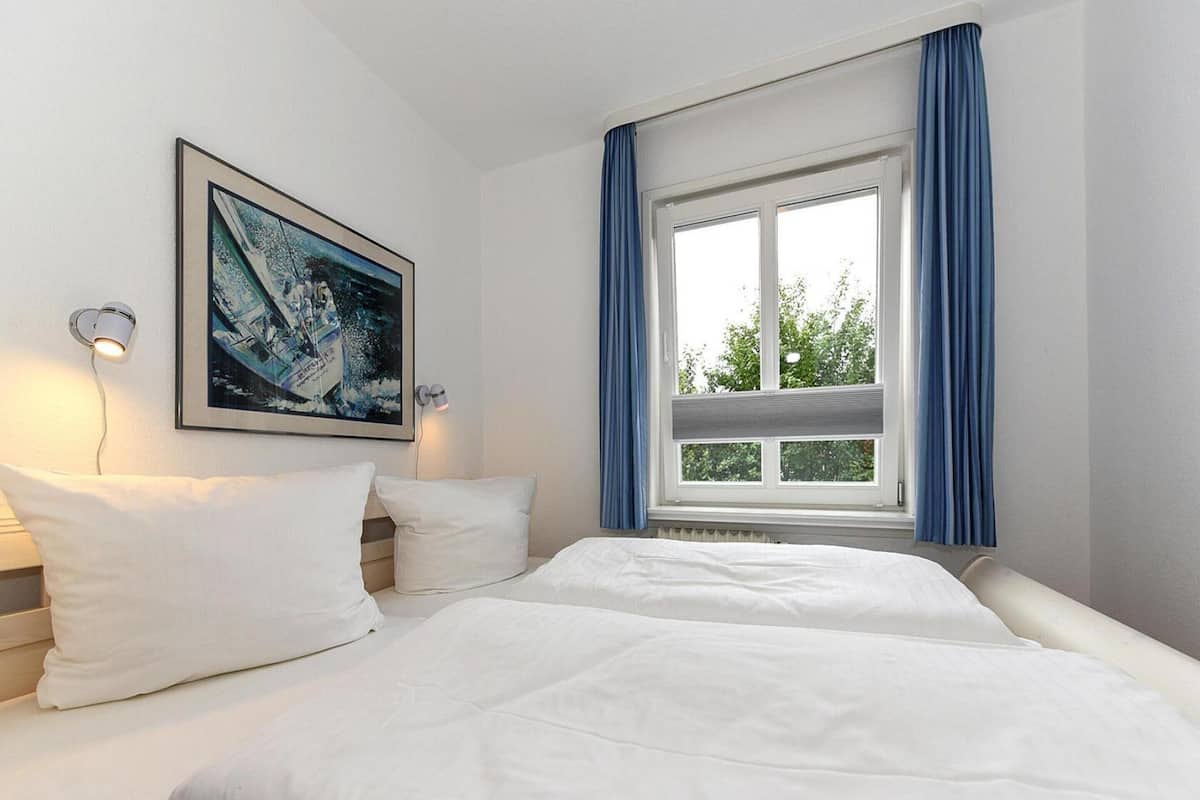 Hotellihuone, jossa on suuri sänky, sinisillä verhoilla varustettu ikkuna, seinään kiinnitetty valaisin ja kehystetty taulu.