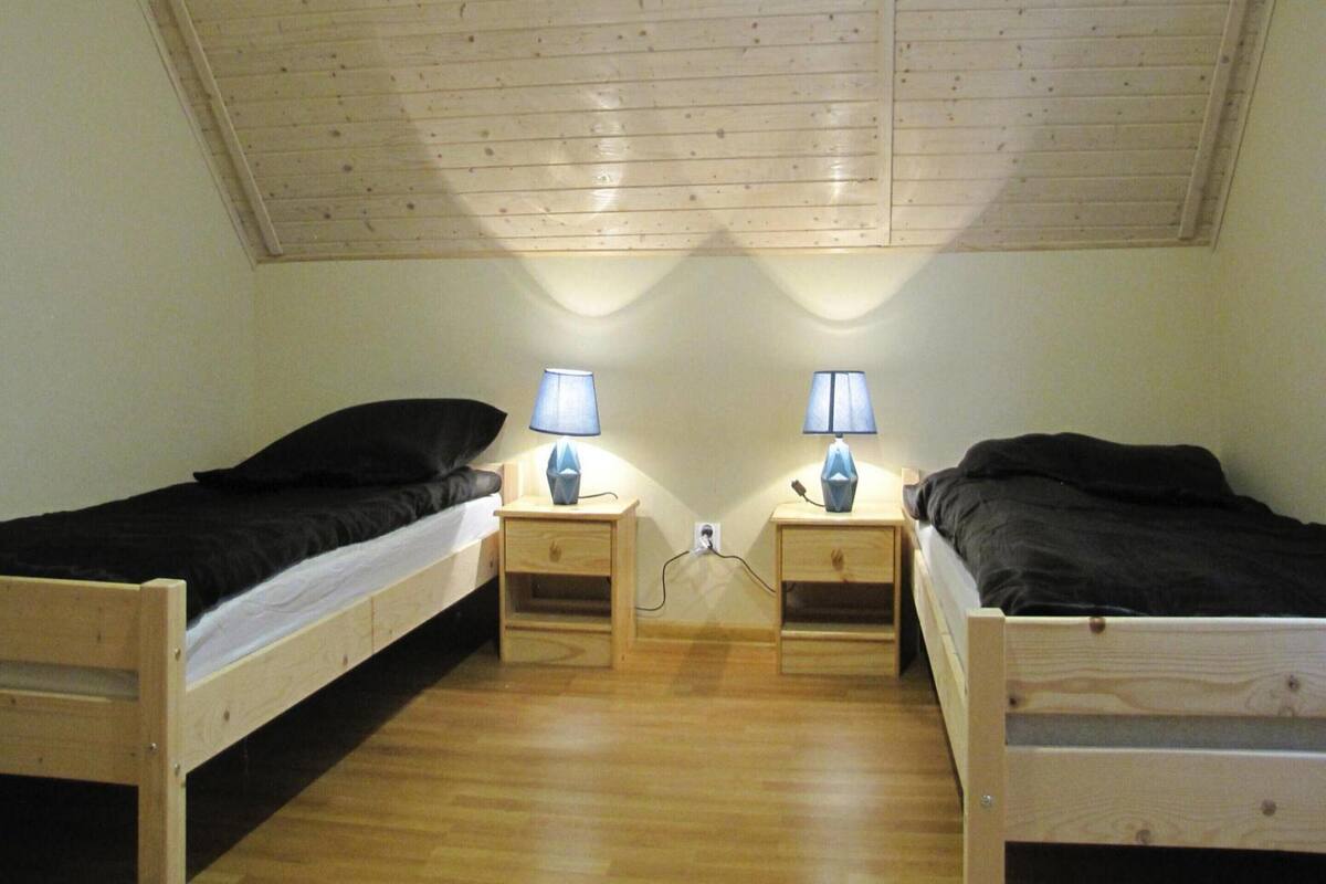A small, cozy room with two wooden beds, each with a pillow and a nightstand with a lamp. The room has a sloped ceiling and wooden flooring.