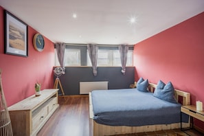 A bedroom with a bed, a bedside table, a lamp, a window, curtains, a radiator, a wall clock, and a framed picture.