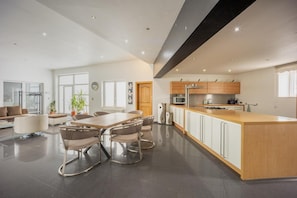 A modern kitchen with a central island, dining area with chairs, and a living space with a sofa and plants.