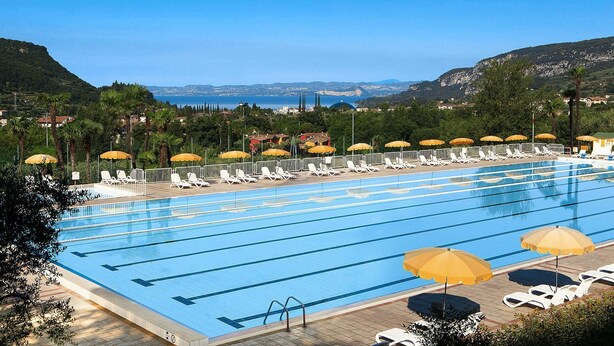 A swimming pool with multiple lanes, sun loungers, and umbrellas, surrounded by greenery and a view of the sea and hills. - Apartments Poiano in Garda With Pool (Garda)
