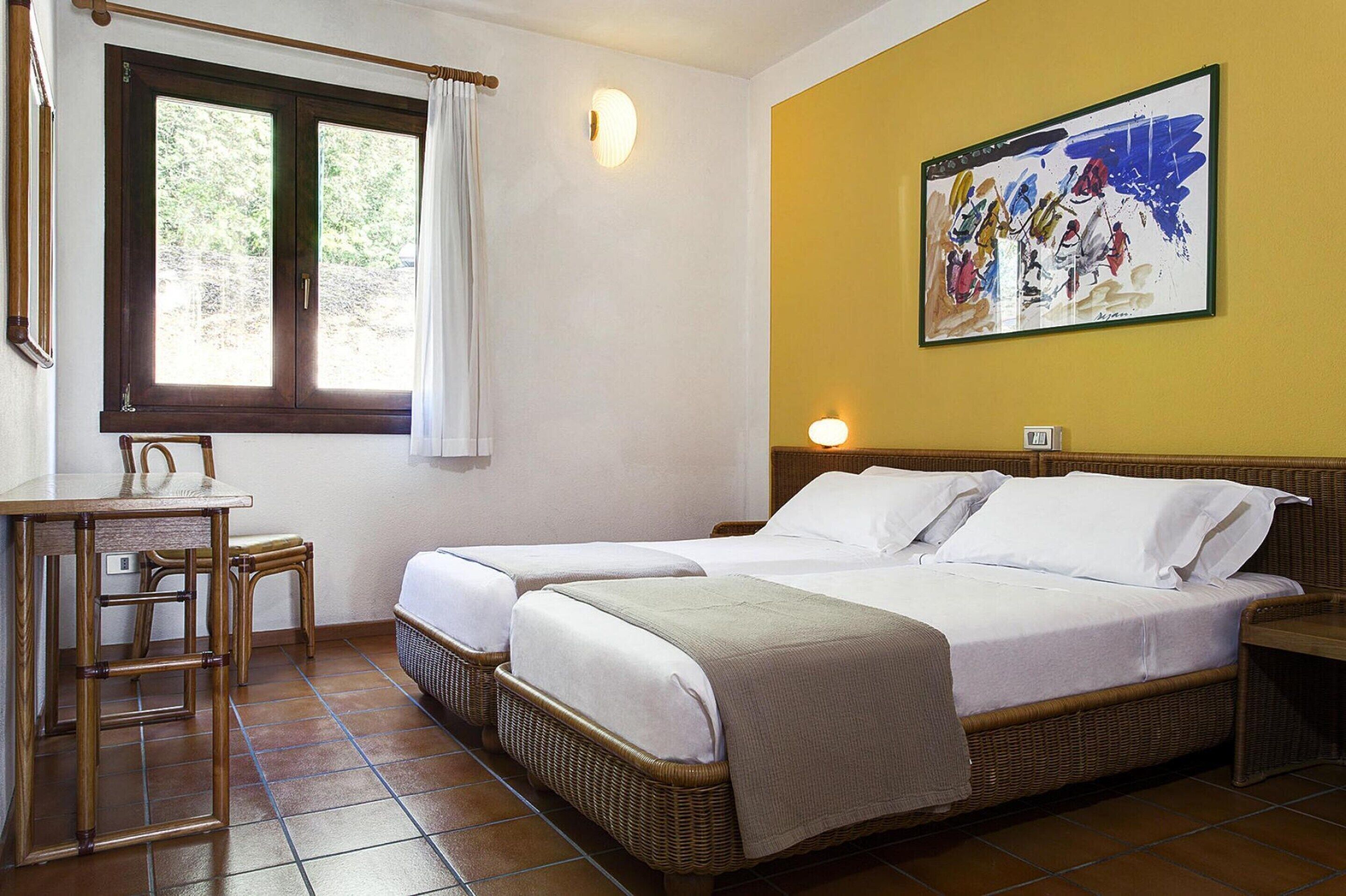 A room with two beds, a dining table, chairs, a window with curtains, and a wall-mounted painting.