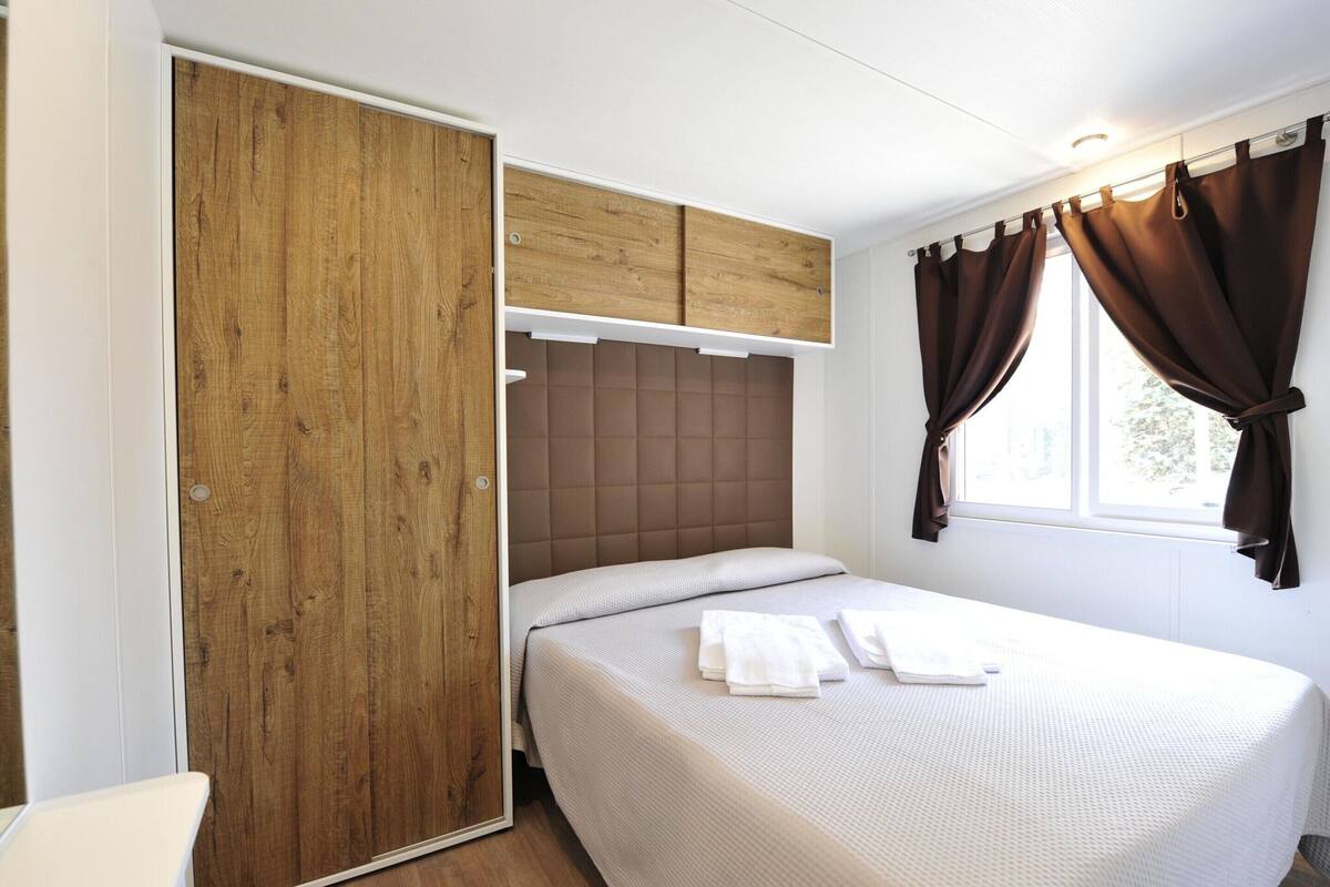 A bedroom with a bed, a wooden wardrobe, a window with curtains, and a headboard.