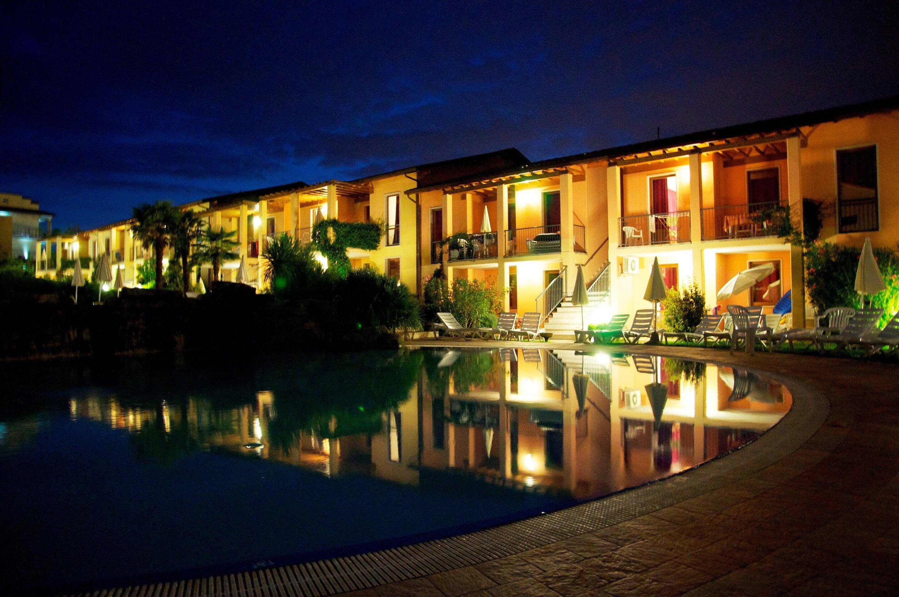 A hotel with a swimming pool, outdoor seating, and a well-lit facade at night.