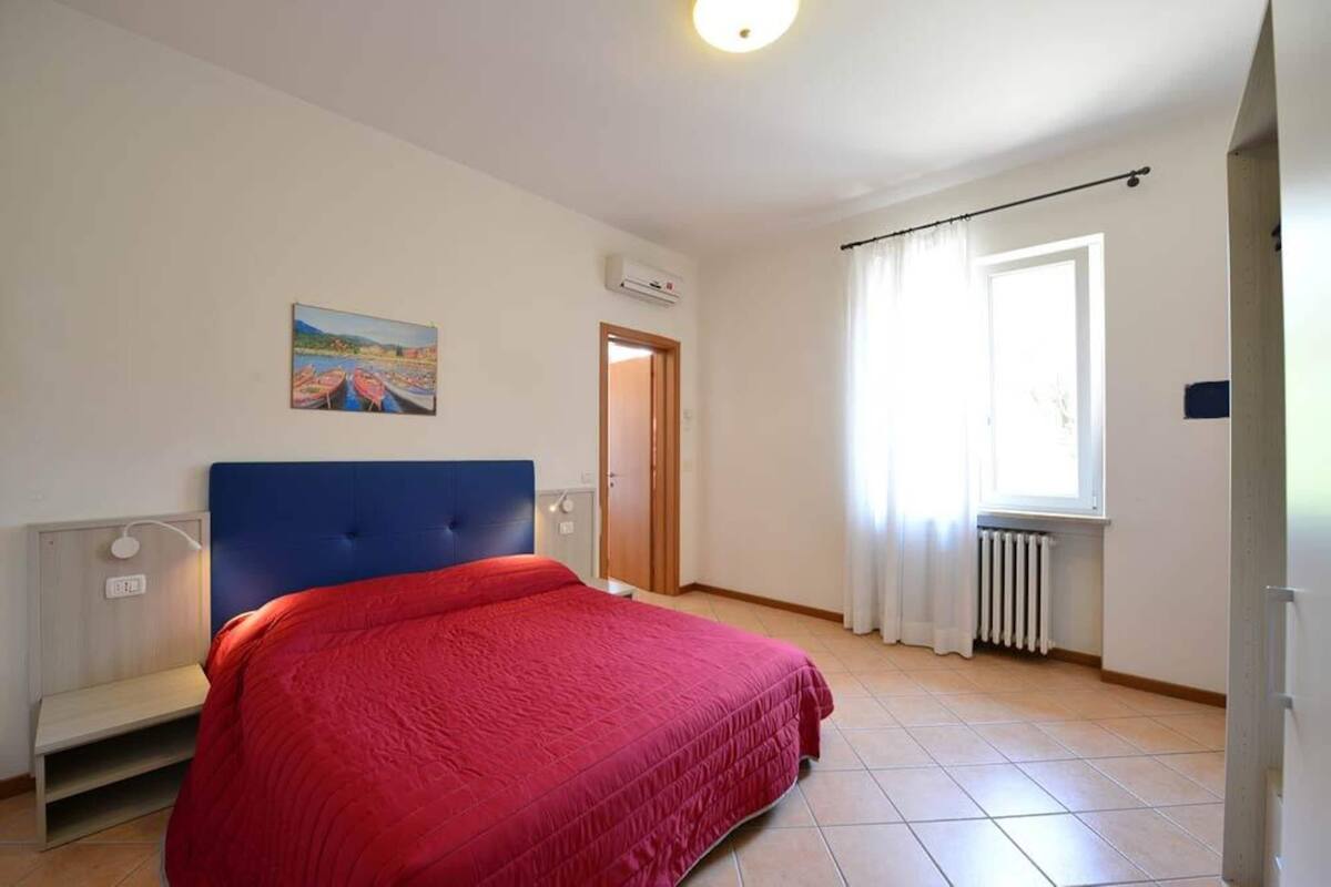 A bedroom with a bed, a painting on the wall, an air conditioning unit, a window with curtains, and a radiator.