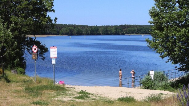 Une vue sur un lac, avec une plage de sable, une rambarde en métal et deux personnes debout au bord de l’eau.