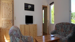 Un salon avec deux fauteuils à motifs, une table basse en bois, un téléviseur à écran plat posé sur un meuble en bois, et une porte donnant sur l’extérieur.