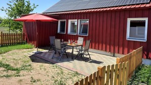 Un bâtiment rouge surmonté d’un toit équipé de panneaux solaires, une terrasse avec une table et des chaises, et une clôture en bois.