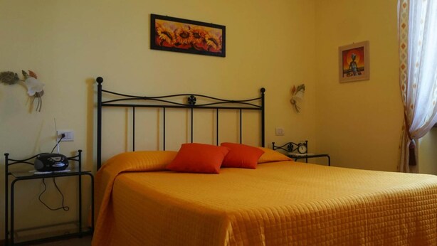 A bed with an orange bedspread and two pillows, a black metal headboard, a wall-mounted painting of sunflowers, and a small table with a radio. - Apartments Casa Tamas, Limone (Tremosine)
