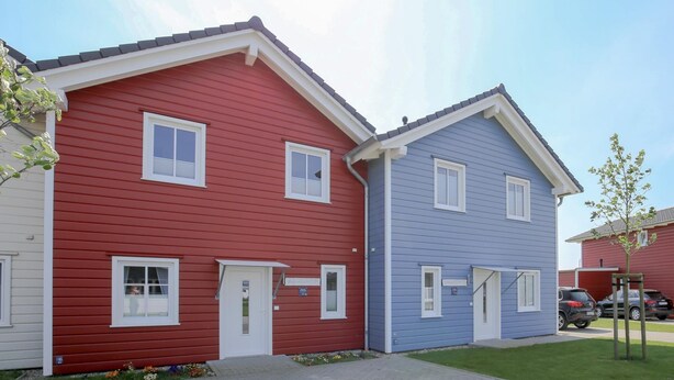 A row of two-story houses with red and blue exteriors, a paved driveway, and a small tree in the foreground. - Exclusively Furnished 5-star Holiday Home Near the Beach (Dagebuell)