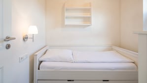 A small, white, single-bed room with a bedside lamp, a wall-mounted shelf, and a door with a handle. - Exclusively Furnished 5-star Holiday Home Near the Beach (Dagebuell)
