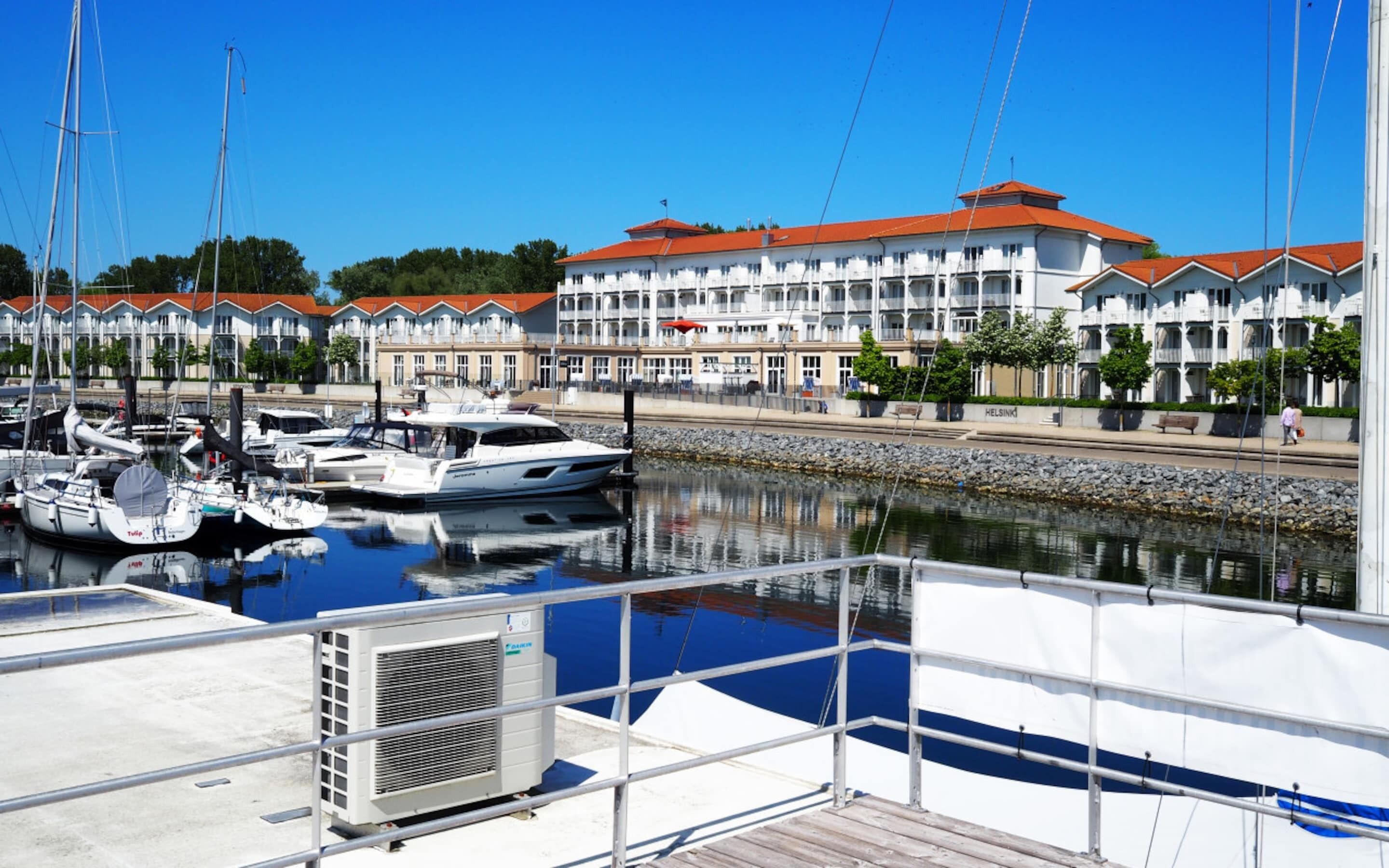 A marina with boats docked, a large building with balconies, and a clear sky.